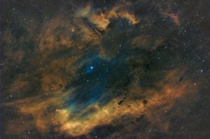 Cette photographie de nébuleuse montre des nuages de gaz interstellaires dans une palette de couleur jaune orangée verte et bleue. Elle semble renfermer une étoile qui figure une perle en son centre