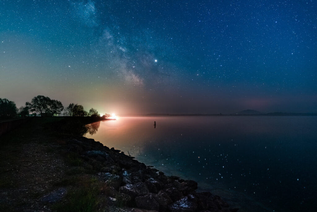 Voie lactée et reflets sur un lac avec au fond un éclairage publique